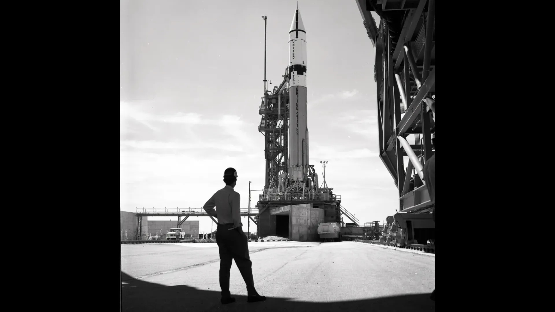 A man stands in front of a rocket on the launch pad in black and white.