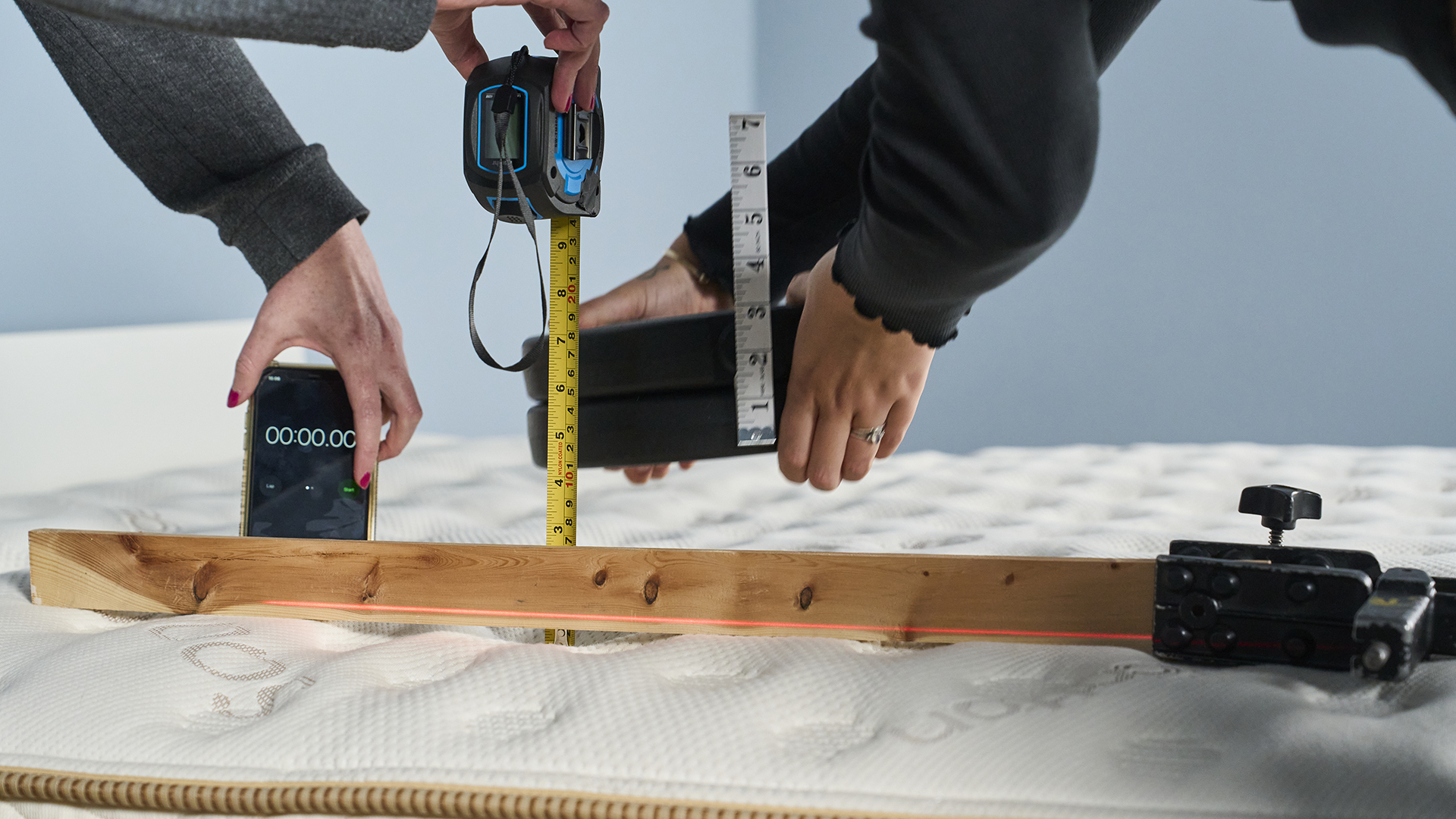 Image shows two members of our reviews team setting up a pressure relief test on the Saatva Classic. One of them holds a yellow digital tape measure and a stopwatch displayed on an iPhone, while the other holds a 10kg stack of weights ready to set them down on the Saatva classic to see how much the weight indents. This gives us an objective measurement of how much the mattress will contour to a weight. The stopwatch will time the weight sinking in for five minutes, then at the end of the weighted indentation test it will be used to time how long the Saatva Classic's materials take to recover. During all of our tests the Saatva Classic recovered instantly, demonstrating best-in-class responsiveness. That makes it an excellent mattress to move around on, and a bed that prioritizes excellent support and contouring comfort.