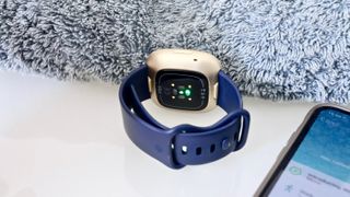 En Fitbit Versa 3 med ett blått armband som ligger på ett ljust skrivbord, med bandet vänt mot kameran.