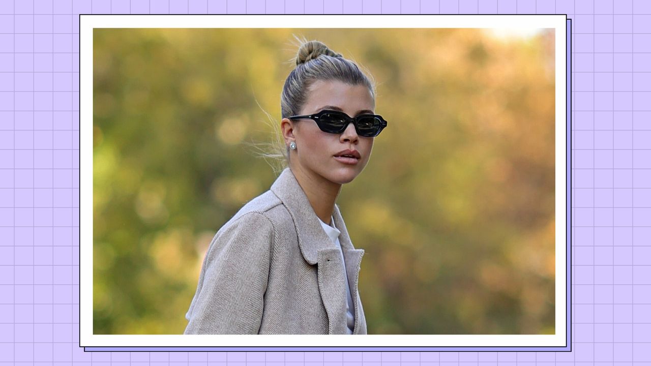 Sofia Richie pictured with her hair up in a bun and wearing a cream coat and black sunglasses/ in a purple template