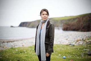 Julie Graham as Rhona Kelly in Shetland, standing on grassland with the beach and the sea in the background. She is wearing a long grey knitted scarf that runs down to her waist, over an open wax jacket and a blue top, with a pair of dark trousers