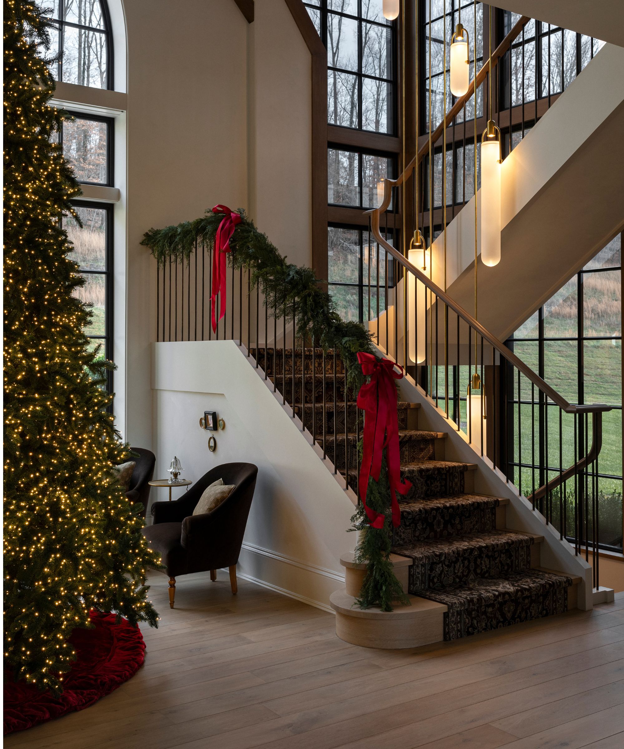 modern home with large staircase wrapped in a a foliage garland with red bows with a giant tree just styled with warm white lights and a red velvet tree skirt
