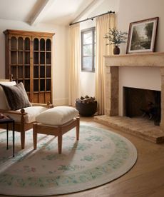 A white armchair and footrest on a cream and green circular rug; in the background, a case of flowers and a landscape picture on a mantel above a fireplace; an antique dresser against a white wall; and sheer curtains around a window. 