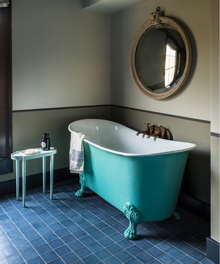 A bathroom with dark blue floor tiles, a turquoise bath and a round wall mirror