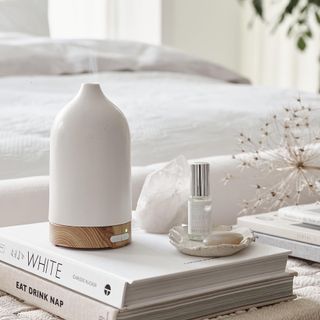 The White Company electronic diffuser sitting on top of a white book and a eat drink nap book, with a fragrance oil and crystal next to it in a white bedroom