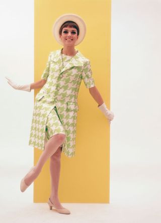 A woman modelling women's fashions in a studio portrait, wearing an enlarged green and white dog-tooth check two-piece outfit, and a straw hat, circa 1960