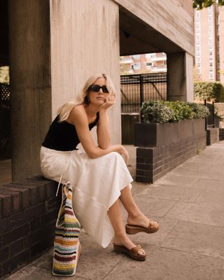 British fashion influencer Lucy Williams sits outside in a one-shoulder black tank top, white maxi skirt, clog slide sandals, and a colorful knitted tote bag