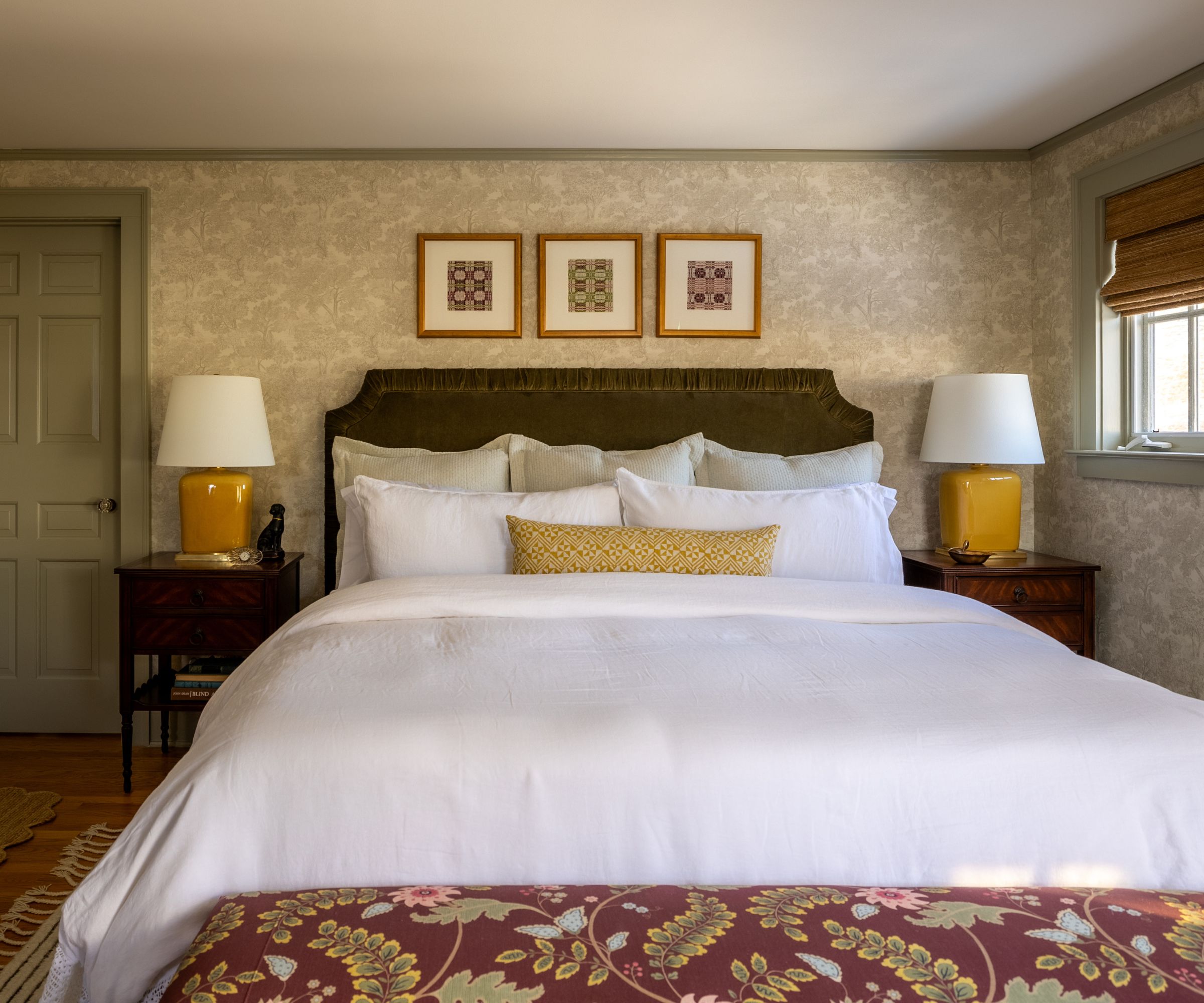earthy cozy bedroom with soft green floral wallpaper with dark green velvet ruffled headboard with vintage bedside tables with yellow lamps