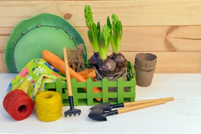 A wooden table with a display of gardening gifts, including a green gardening hat, garden hand tools, green and yellow twine, bulbs and compostable plant pots