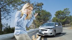 A frustrated-looking woman stands next to her wrecked car and talks on her smartphone.