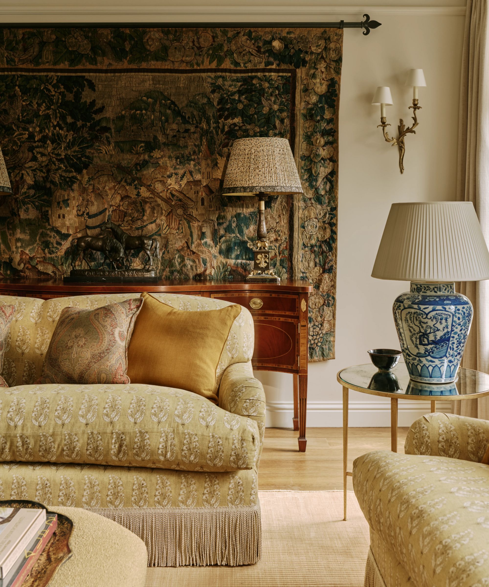 a warm neutral living room in london with golden toned floral sofas with fringe trims, antique furniture, and a vintage tapestry on the wall