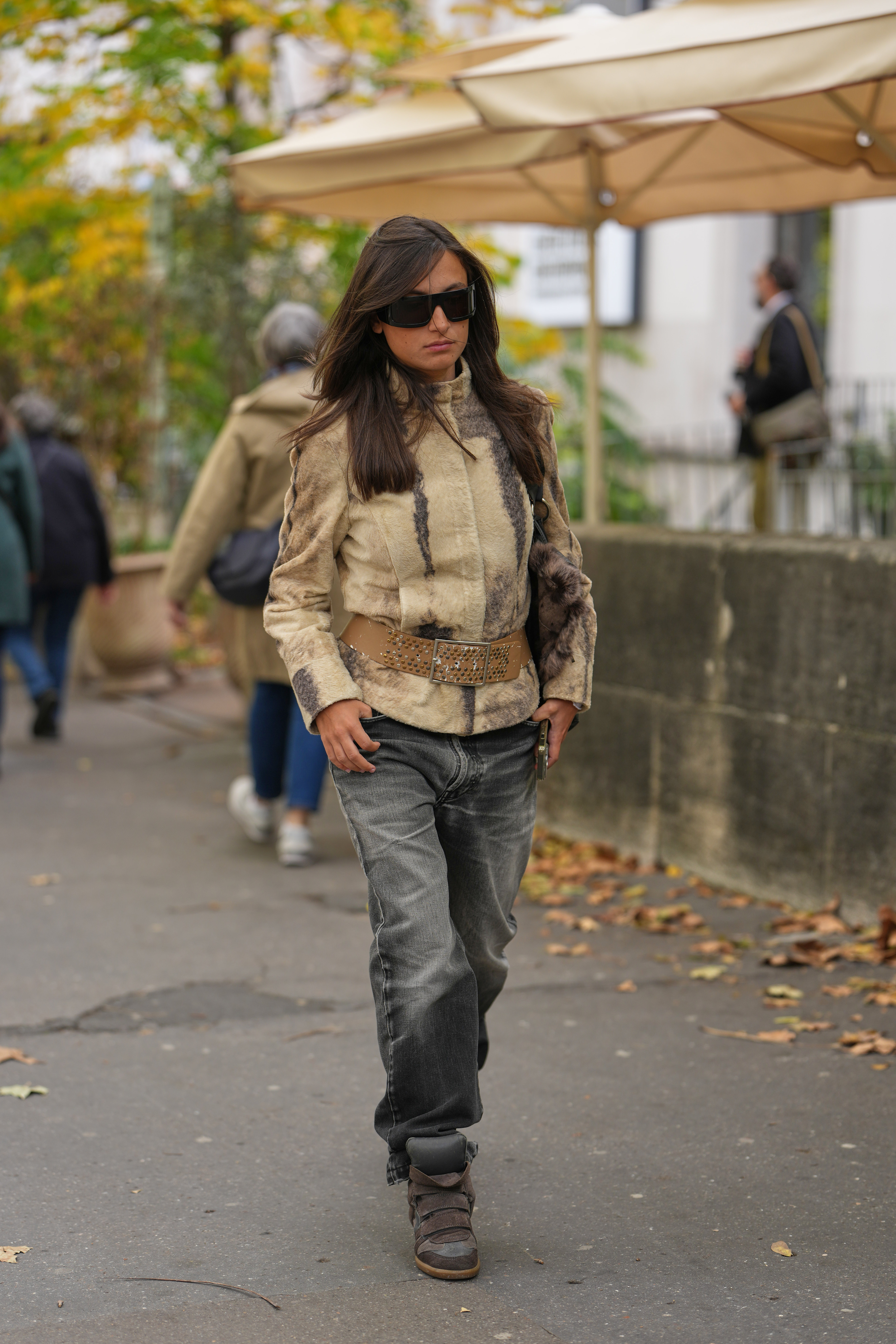 PARIS, FRANCE - OCTOBER 05: A guest wears long straight dark brown hair with a slight center part, large rectangular black futurist sunglasses, a brown fur clutch bag, a beige shearling jacket with darker brown vertical markings and a high collar, a wide tan leather belt with punched studs and a large rectangular metal buckle, faded grey-black relaxed-fit denim jeans, brown suede wedge sneakers shoes with velcro straps and rubber soles from Isabel Marant, outside Akris, during Paris Fashion Week - Womenswear Spring Summer 2026, on October 05, 2025 in Paris, France (Photo by Edward Berthelot/Getty Images)