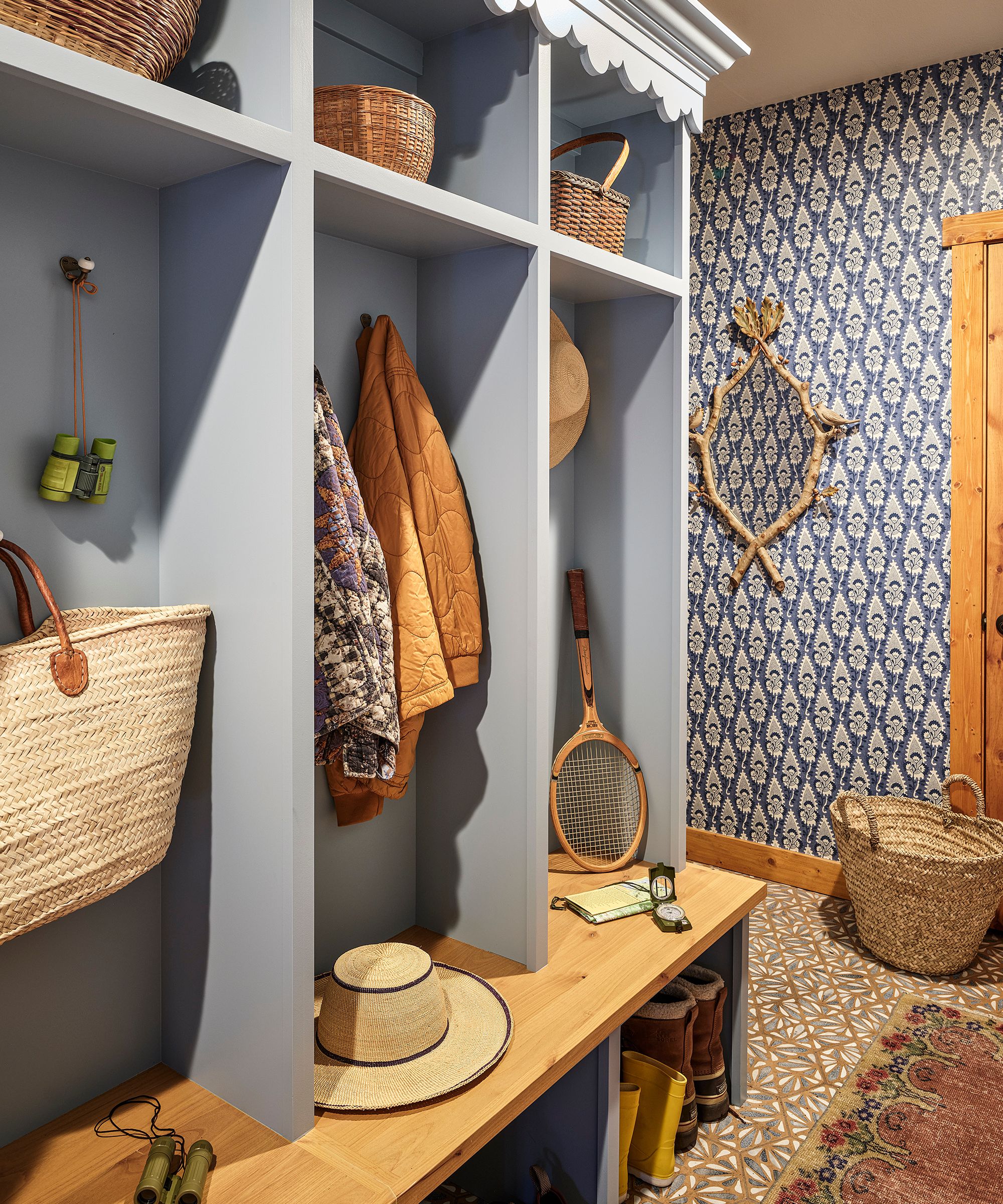 a pale blue mudroom with block print floral wallpaper, tiled floor, and a built-in wall of storage with a bench and hooks for bags and bags