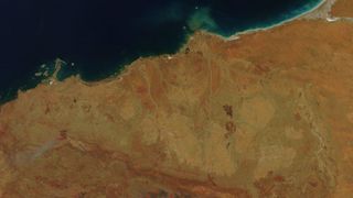 Satellite image of the Pilbara Craton in Western Australia shows a yellow and red coastline as seen from above next to a deep-blue ocean