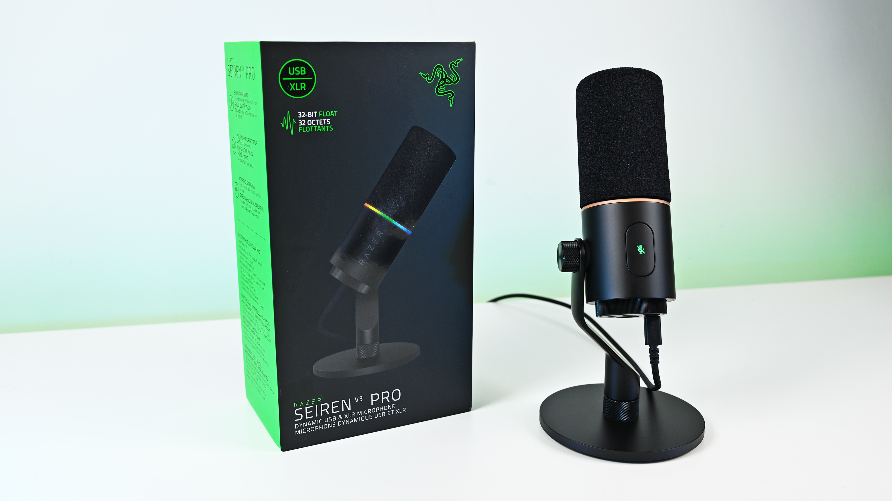 Angled shot of the Razer Seiren V3 Pro microphone on a white desk next to its packaging, highlighting the green side panel of the box.