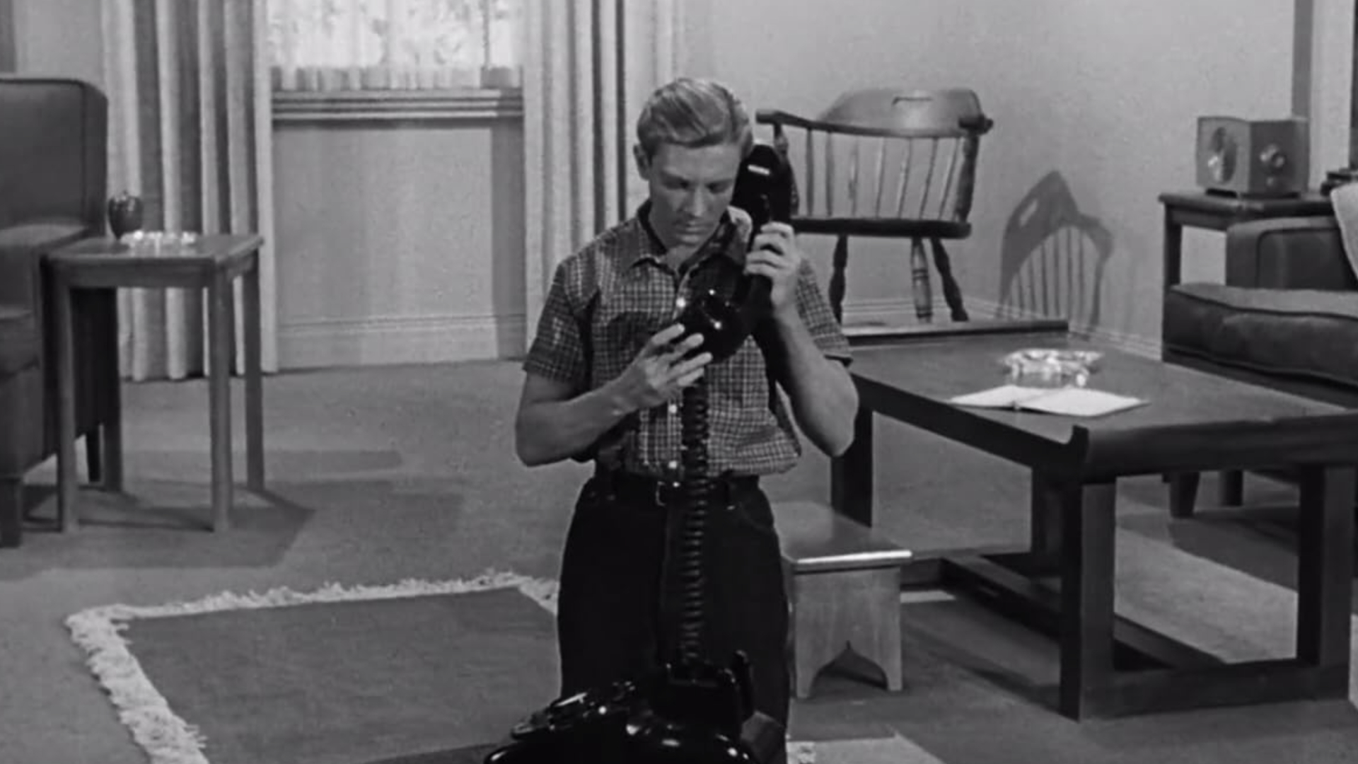 A man, who is the same size as the livingroom furniture in the room, holds an oversized house phone