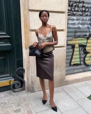 Influencer wears a brown leather knee length skirt, black pointed toe heels, a silk camisole, and a black clutch.