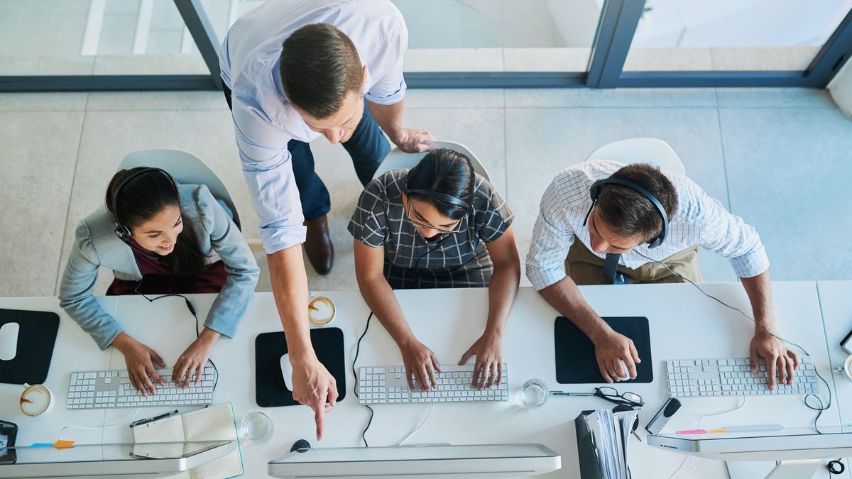 A top-down shot of a manager instructing three IT workers, who are wearing headsets and sat at a desk facing a bank of monitors.