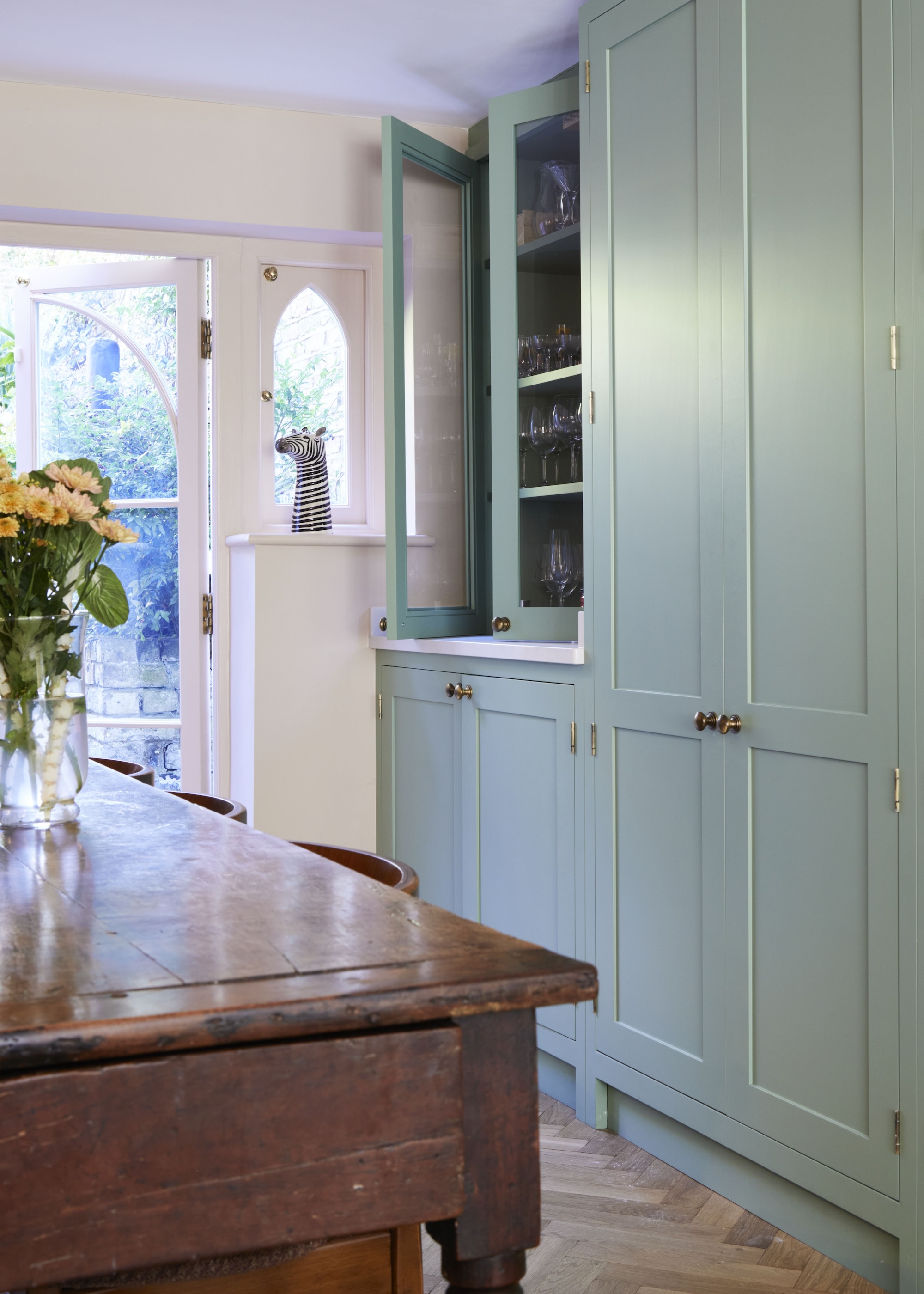 A glass fronted cabinet is opened slightly ajar with a view of the dining table that has some plants in a vase with water inside. There is also a view of the other kitchen cabinets that remain closed