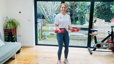 Maddy Biddulph performs a biceps hold in her living room at home. She is standing and smiling at the camera, with her elbows bent and held close to her side. She grasps a pair of dumbbells in her hands. Behind her we see sliding glass doors leading to a garden and an exercise bike.