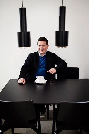 A caucasian man sitting on a black table with 4 black chairs against a white wall with white mug, on a white saucer with silver teaspoon in it placed in front of him on the table. Above the table are 2 hanging black ceiling lights