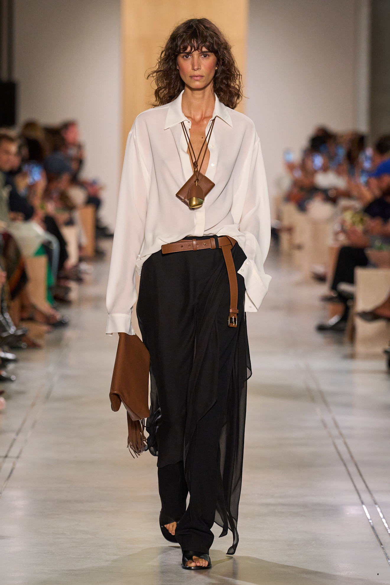Layering staples for spring 2026 is shown in a photo from Michael Kors's runway show, with a model walking while wearing an oversized white button-down tucked into a sheer black skirt layered over black trousers. The rest of the outfit includes two cord necklaces, a tan belt, a tan clutch bag, and black sandals