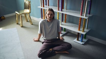 A young man dressed in a gray T-shirt and burgundy trousers sits with his legs crossed on the floor of a dimly lit room containing large-scale colorful trophies and a zig-zag-cut, golden chair.