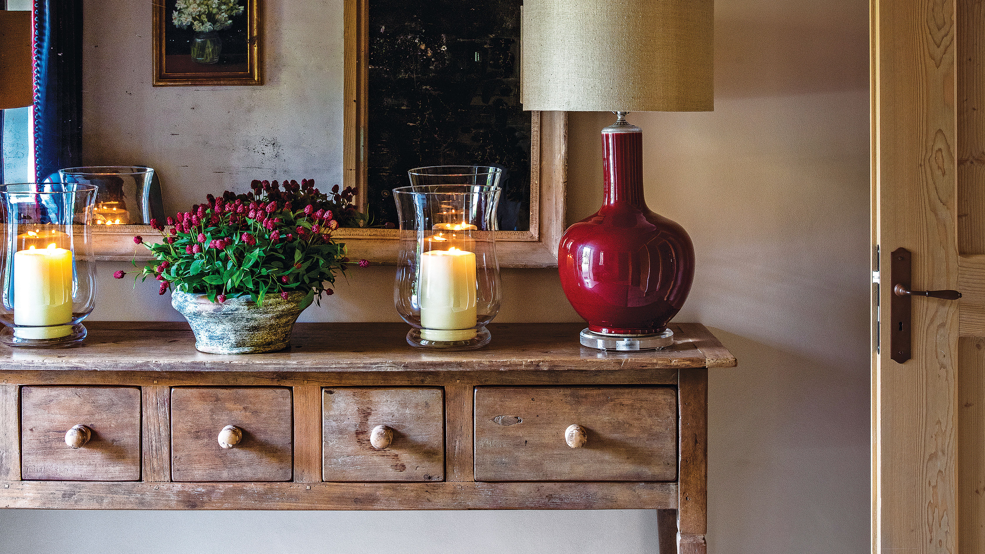 Country-style wood sideboard with red lamp, candle in a glass vase, bowl of red flowers in a marble pot and a mirror 