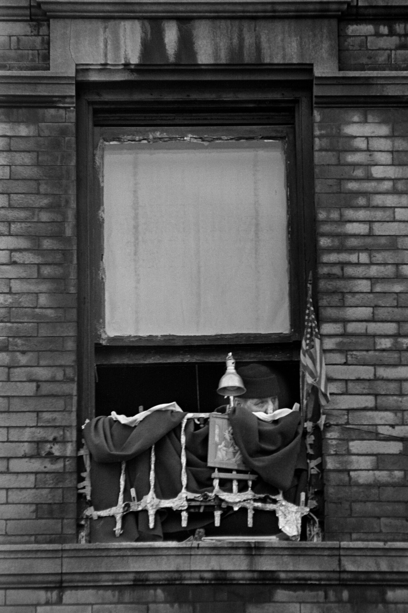 A person in a dark woolly hat peers out from an apartment window draped with fabric and foil, with a small lamp, a framed picture and an American flag at the sill.