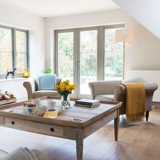 neutral living room with wooden floor, glass doors and window, matching light brown armchairs and a large square coffee table