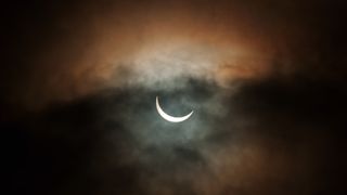 partially eclipses sun appears as a thin crescent behind clouds.