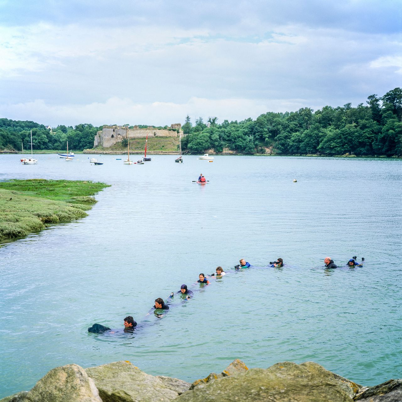 A group of people wearing wetsuits form a line in shallow water during a training exercise in a sheltered bay.
