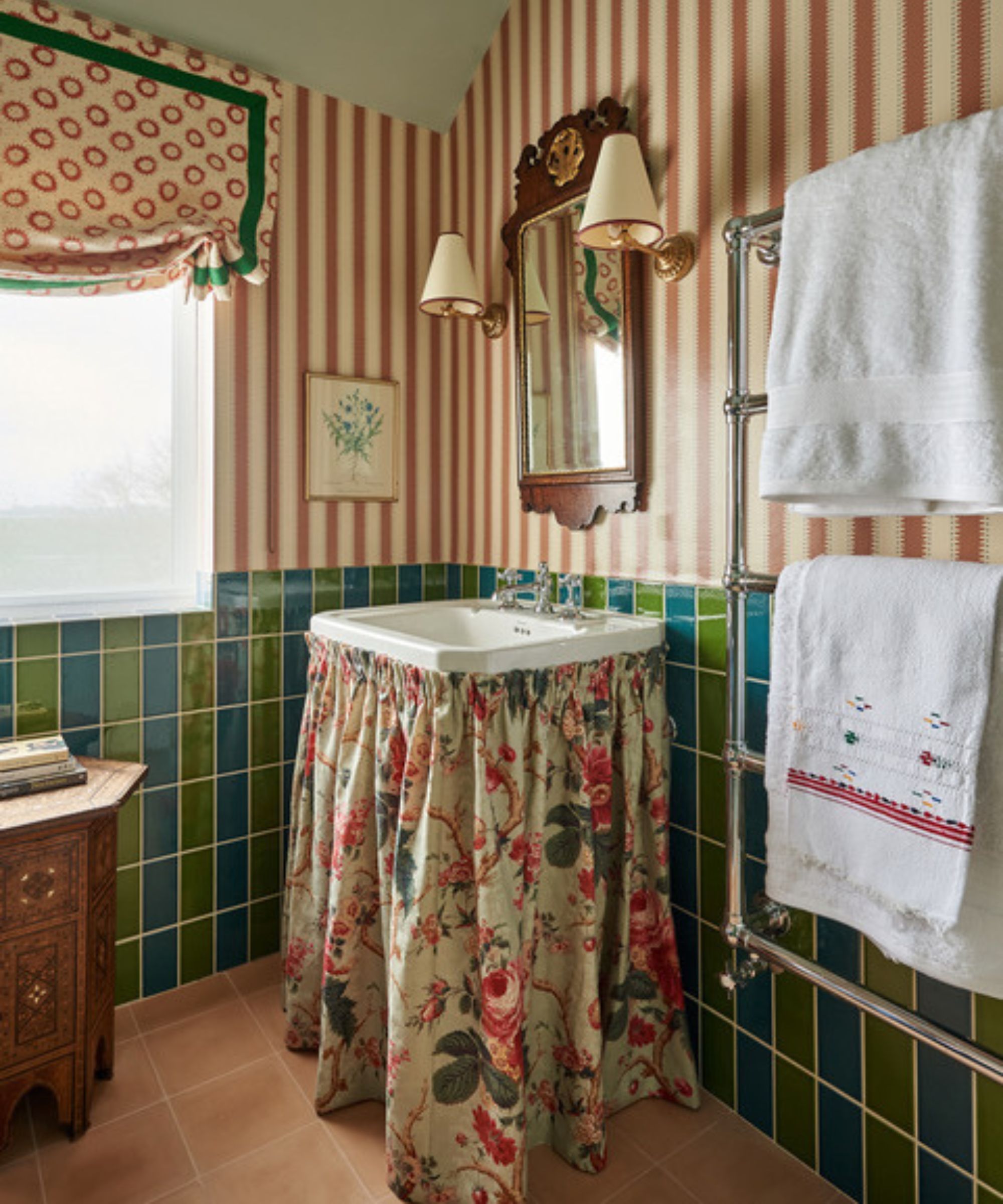 A maximalist bathroom with blue and green checkerboard wall tiles, striped wallpaper, and a floral vanity skirt