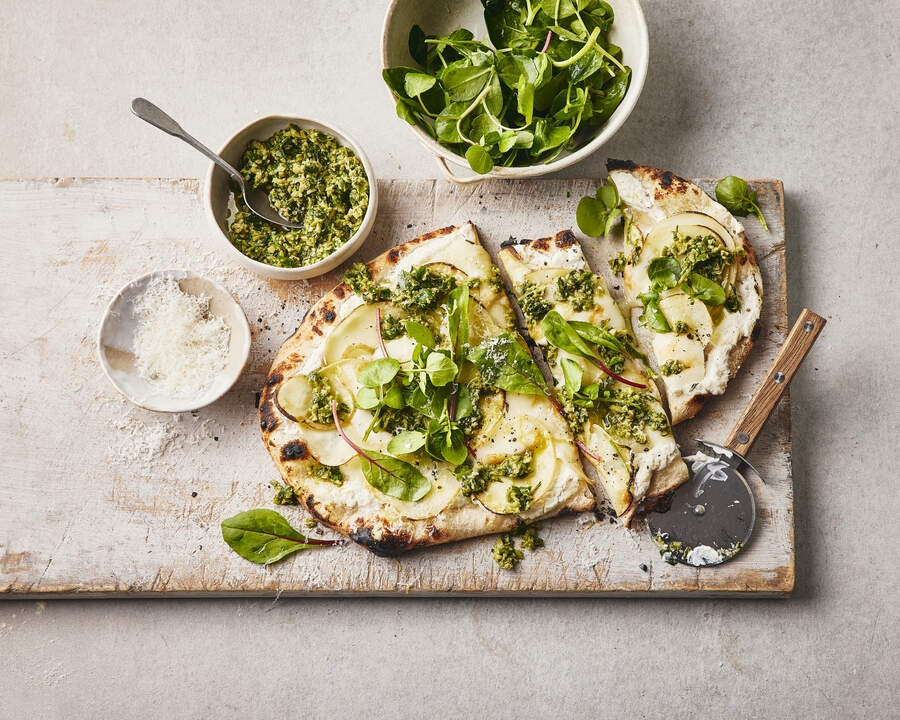 A freshly baked pizza topped with cheese, pesto, and fresh greens, served alongside bowls of pesto and salad.