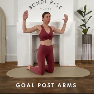 Lottie Anderson performs goal post arms Pilates move at home on a rounded workout mat. SHe is in a living room with a fireplace behind her and a potted plant nearby. She is kneeling, with her arms out to the side, elbows bent at 90-degree angles, hands pointing upwards.