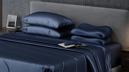 A bed with blue silky sheets, a book, an eye mask, stacked ergonomic pillows, and a gray headboard