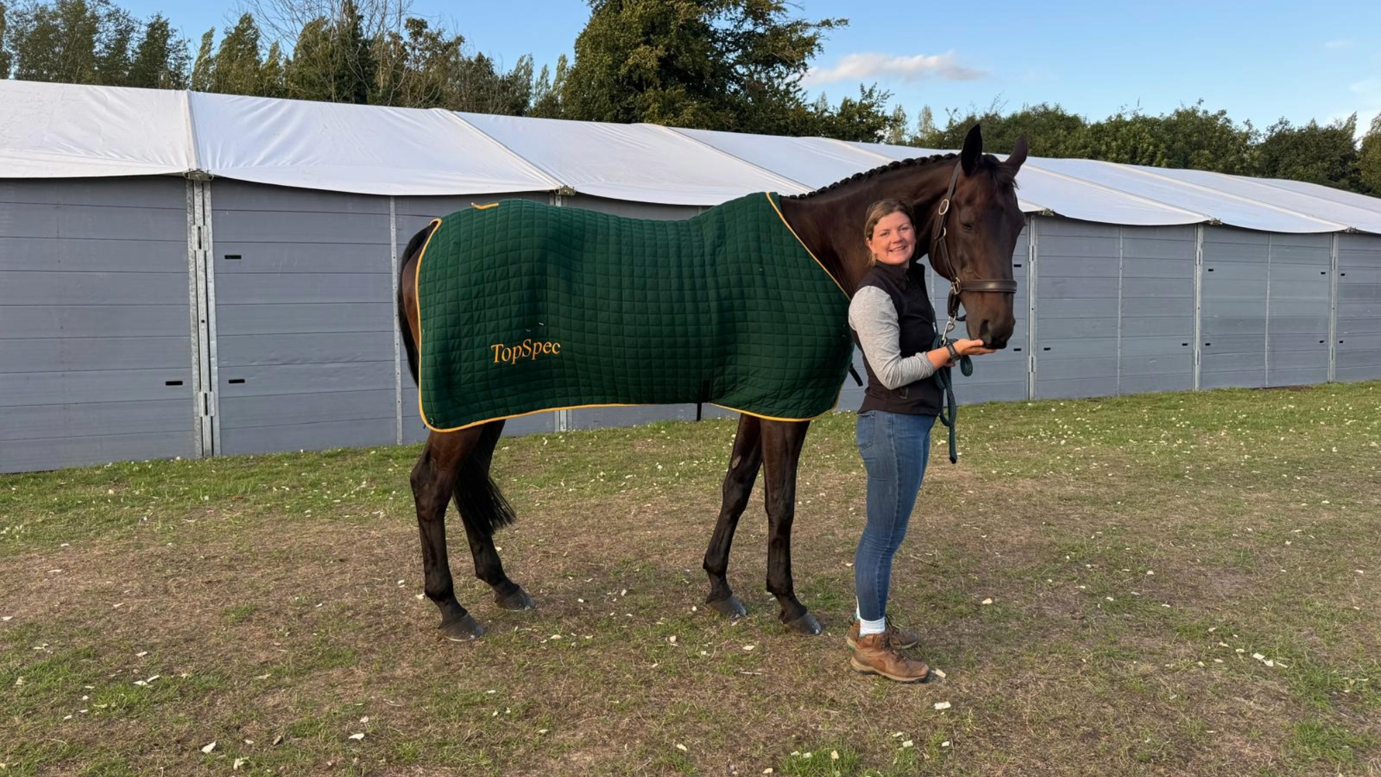 Groom Ruth Asquith stands holding Katie Magee&rsquo;s 2026 Burghley horse Treworra in a headcollar and a TopSpec sponsored green rug