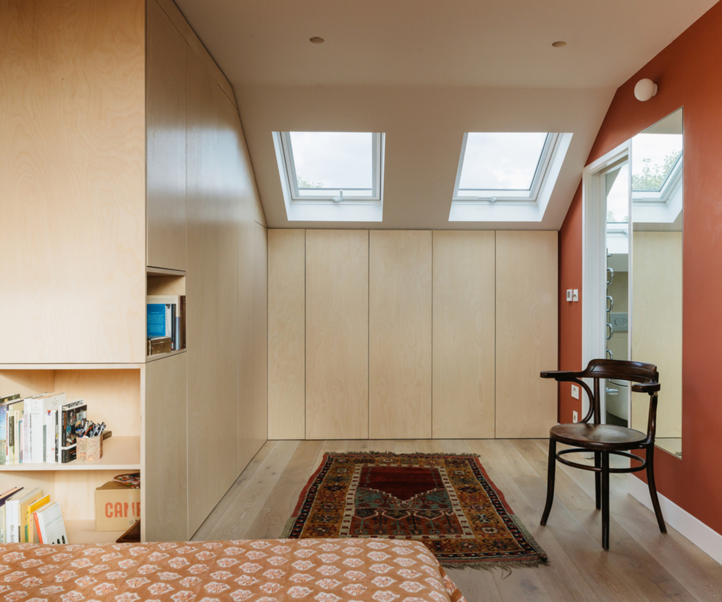 dormer loft conversion bedroom with walls clad in wood with integrated storage, wooden floor, rich red painted wall with large mirror and shelving also recessed into walls, two small rooflights above storage