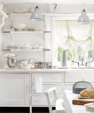White country kitchen, updated units and cupboards with white doors and handles on stilts, butler sink, panelled wall with white shelves containing crockery and glassware, pretty floral curtain in window, grey industrial style pendant lights,