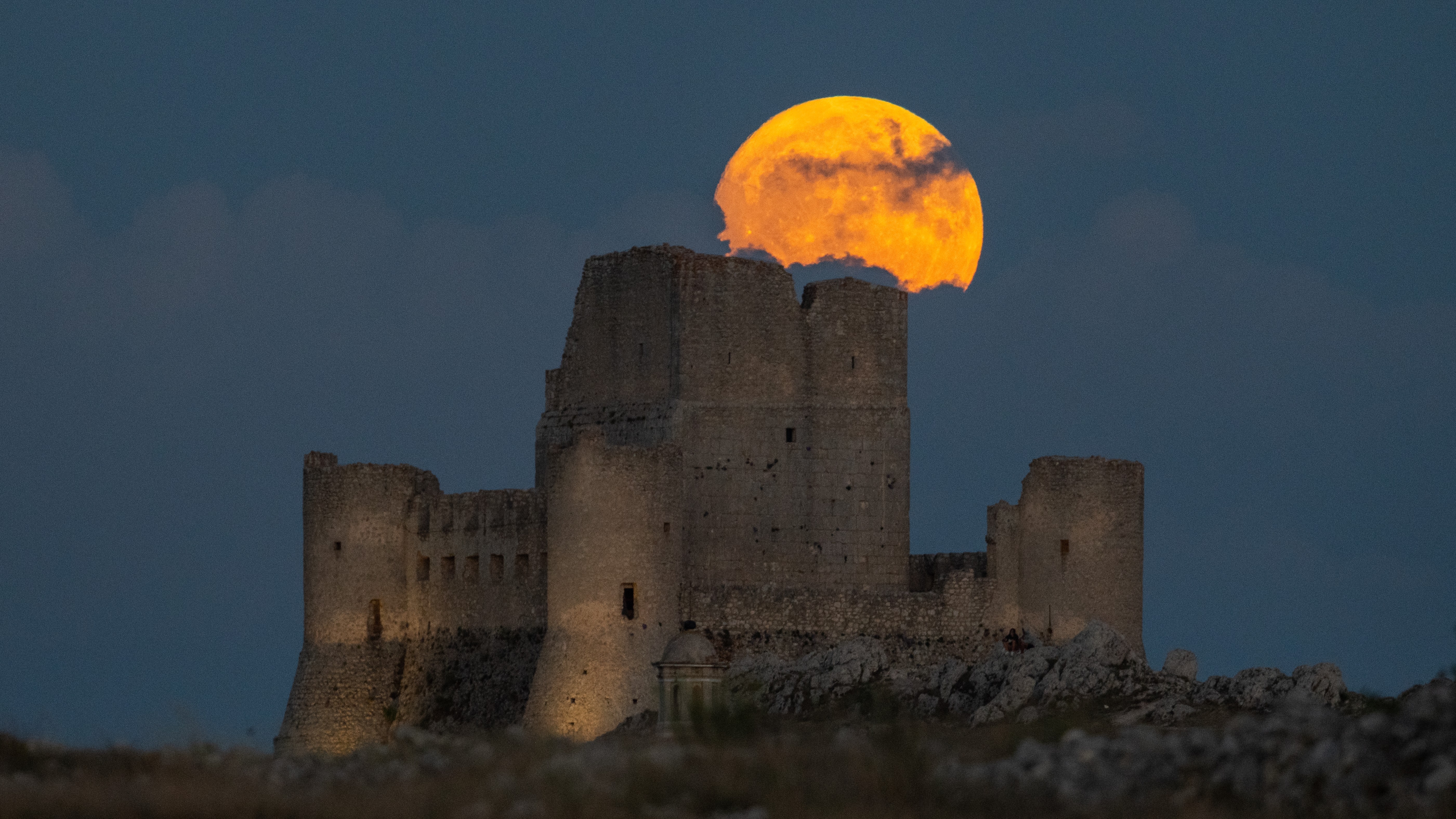 July full moon rising above a castle ruin and appearing very large in comparison to the castle.