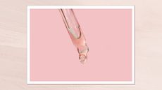 A close up of a glass pipette dripping out a clear serum-like liquid in front of a pink backdrop/ in a textured pale pink template