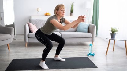 Woman performing a shallow squat on a black exercise mat in a living room setting. there's a sofa behind her.