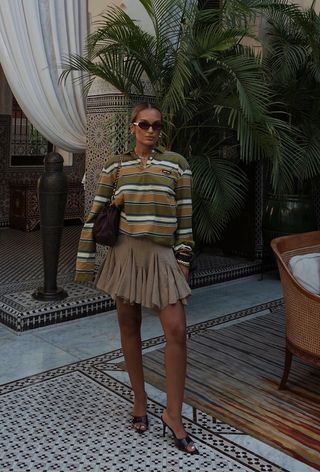 A woman wearing a striped green and brown polo shirt with a pleated tan miniskirt, brown mules, and a brown shoulder bag.