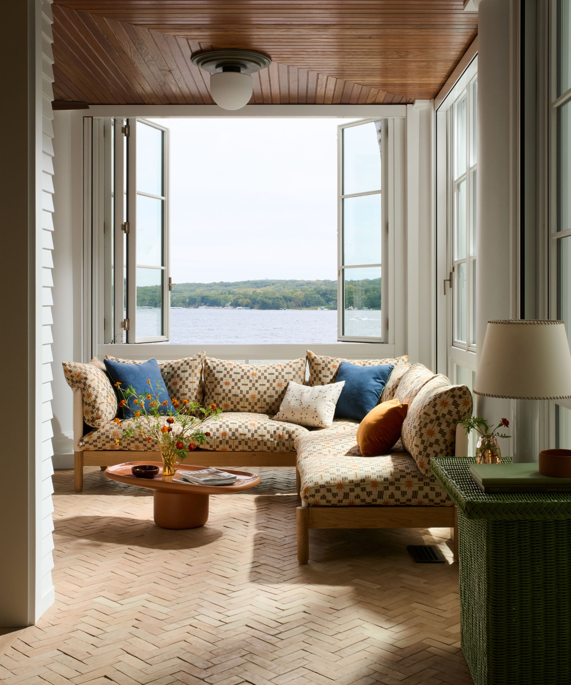 Sunroom with large open window overlooking a lake, exposed brick floor, an upholstered sectional with a geometric print, a round pink coffee table, an antique green wooden console holding a lamp.