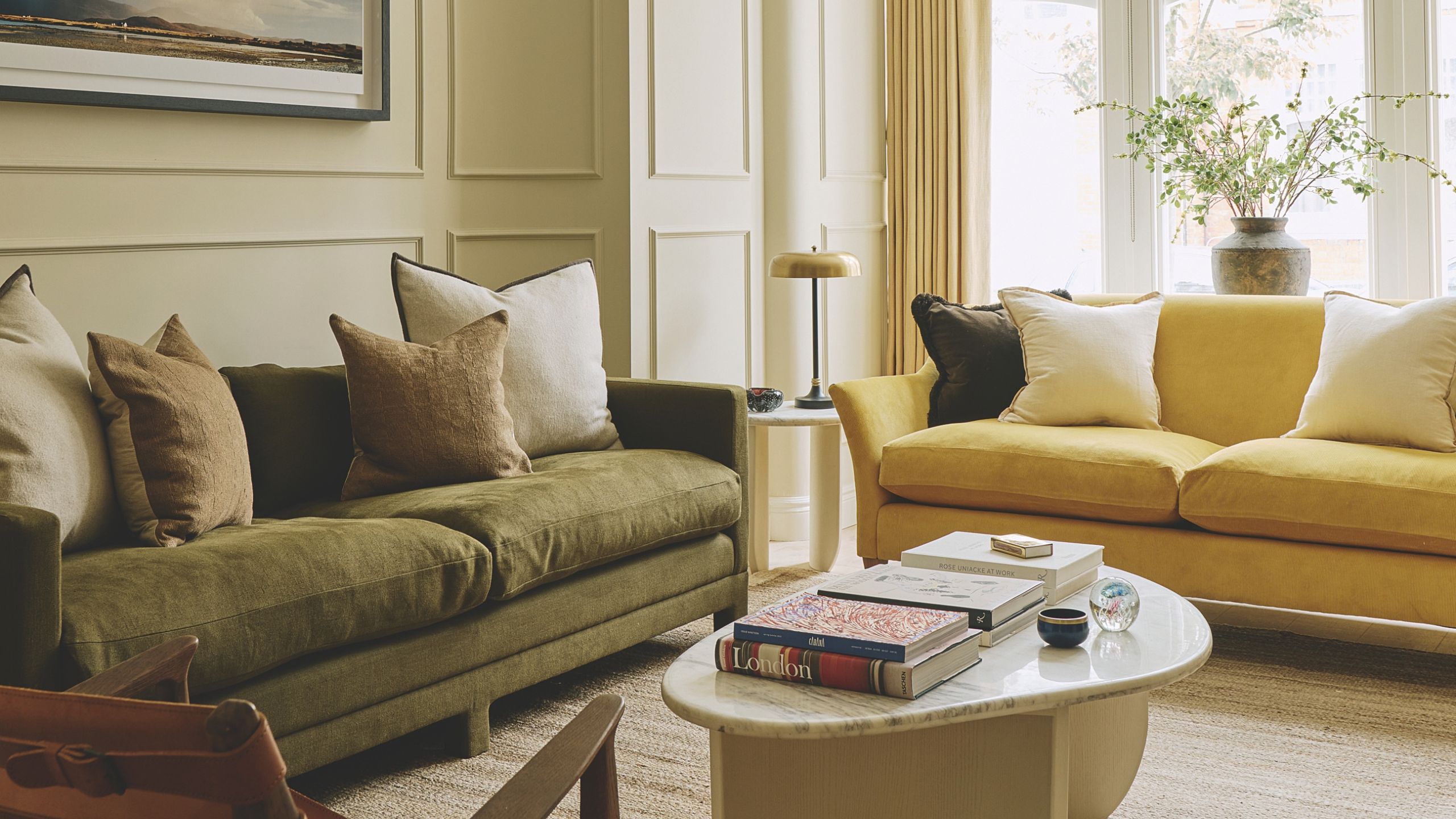 Elegant living room with paneled cream walls, olive green velvet sofa, yellow accent couch, marble coffee table, and large window with curtains