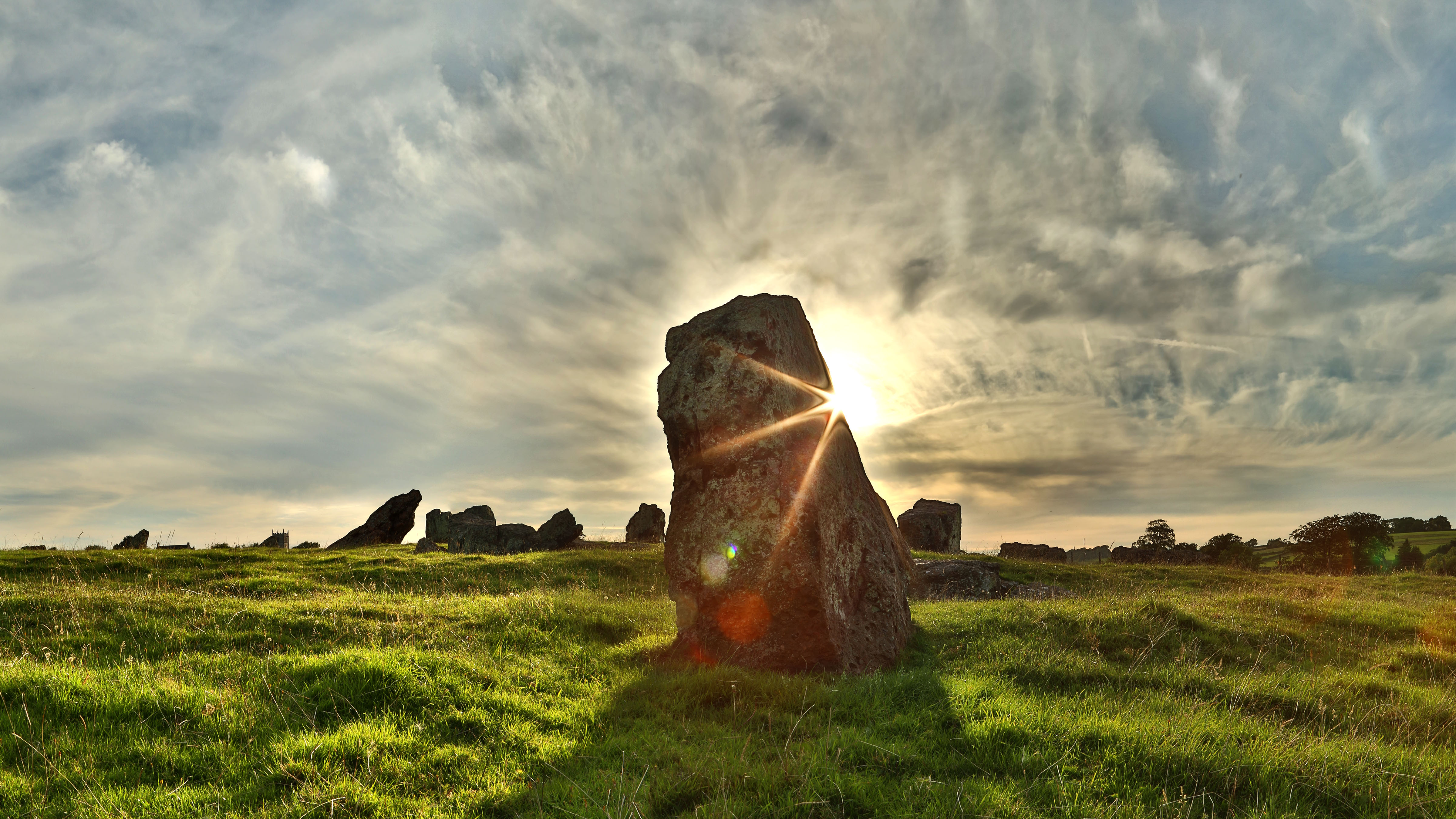 a big stone is in front of the sun's rays. the stone is on grass. Far in the back are other very large stones and trees, and a cloudy sky