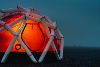 Close up view of an orange tent with a white framework by 66°North and Heimplanet in the evening - the tent is illuminated from the inside and sits on rocky ground outside