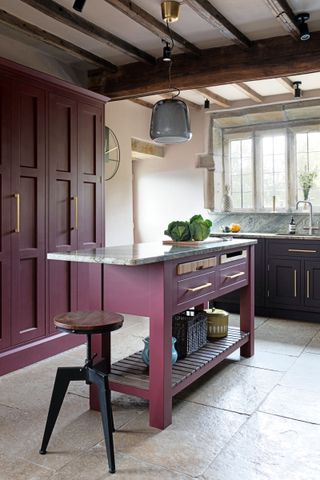 A traditional berry red kitchen with painted kitchen island and cabinetry, pale gray stone flooring and a timbered ceiling.
