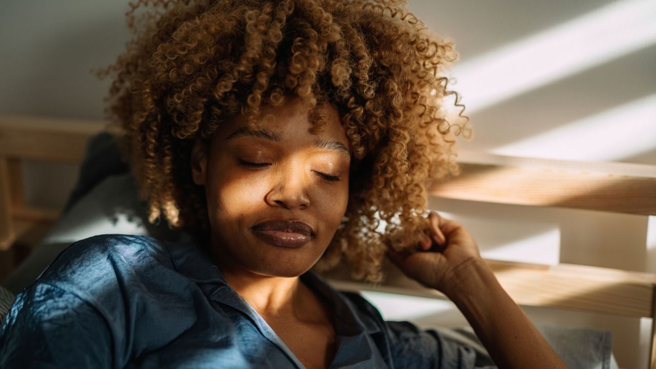 Woman with eyes closed, half lying down in bed, and sun coming through window against her face, practising exercises after learning what is grounding