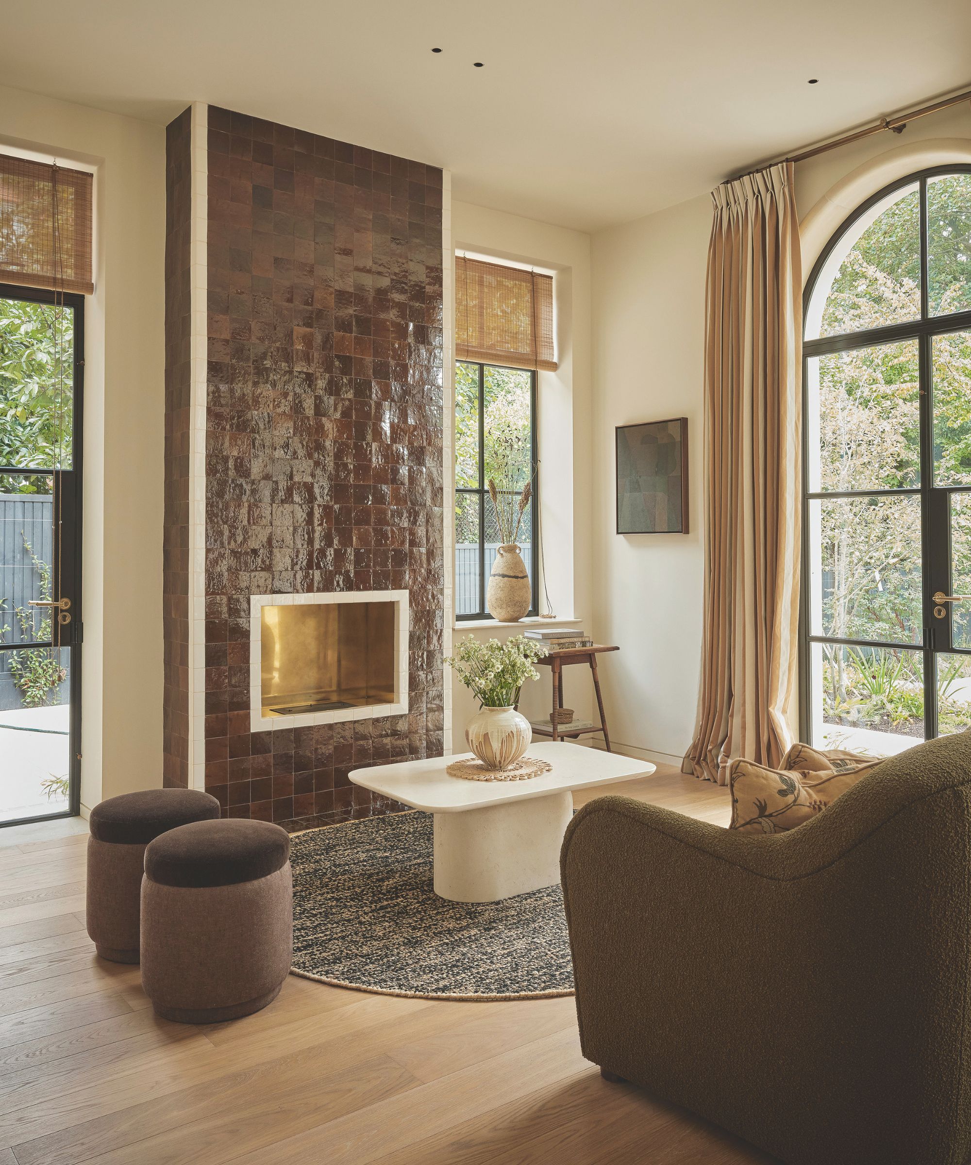 Cream living room with a fireplace that&#039;s tiled in glossy bodwn square tiles. There is a white coffee table on top of a round rug and two brown velvet stools